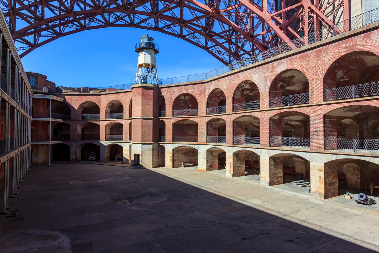 A brick fort with arched walkways and a lighthouse tower in the courtyard, partially shaded, with a large metal structure overhead against a blue sky—one of the most intriguing travel destinations on the West Coast.