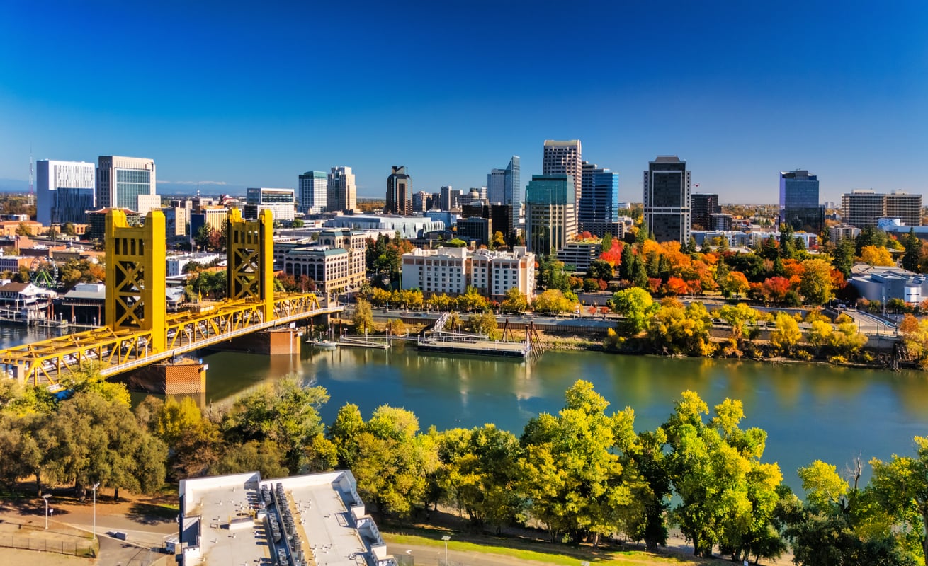 A travel-friendly view of Sacramento’s skyline showcases the iconic yellow Tower Bridge spanning the river, surrounded by trees adorned with green and autumn-colored leaves under a clear blue sky.