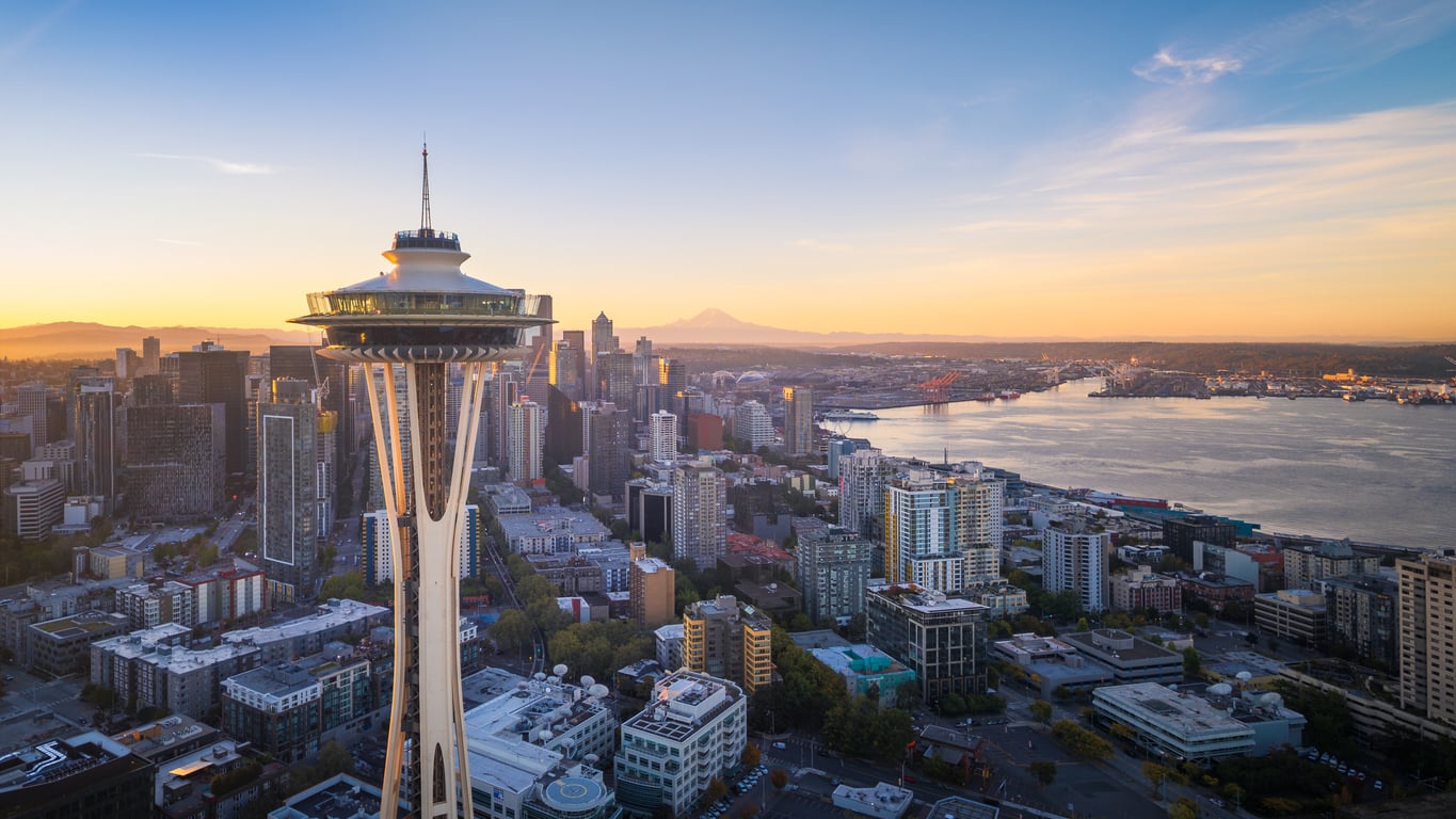 Aerial view of the Seattle skyline at sunset, featuring the Space Needle in the foreground, downtown buildings, and water with Mount Rainier visible in the distance.