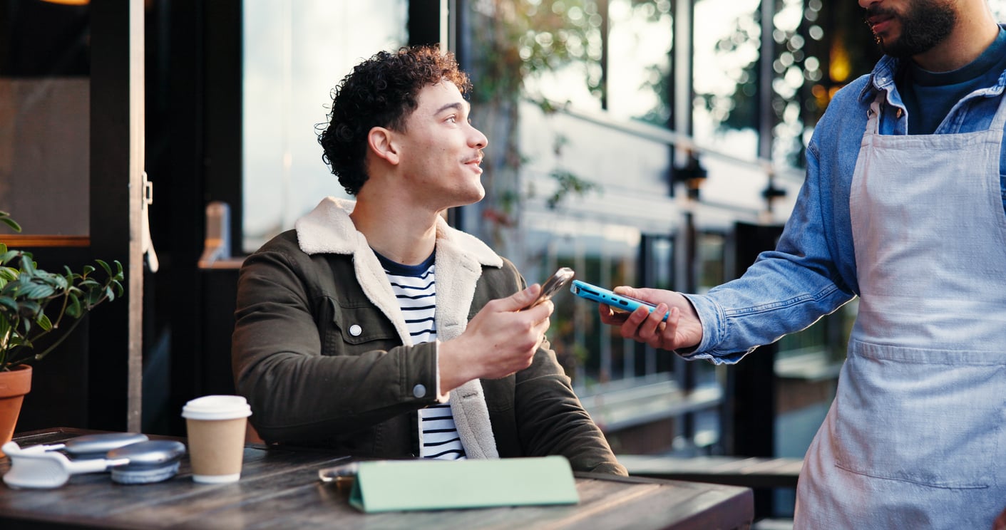 A man, embodying traditional Baby Boomer beliefs in enjoying the simple pleasures of life, sits at an outdoor café and smiles as he pays for his order using his phone, while a server holds a card reader. A coffee cup and small plant are on the table.