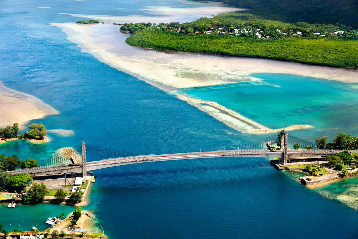 A bridge spans turquoise and deep blue waters, connecting lush green landmasses with sandy shallows visible beneath the surface. Small buildings and vegetation line the shorelines, creating an idyllic scene often featured among top travel destinations.