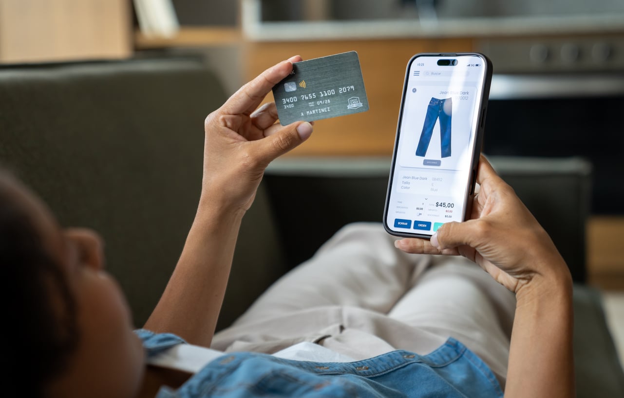 A person lying on a couch holds a credit card in one hand and a smartphone displaying an online shopping page for jeans in the other hand, highlighting how different this experience is from life before smartphones.