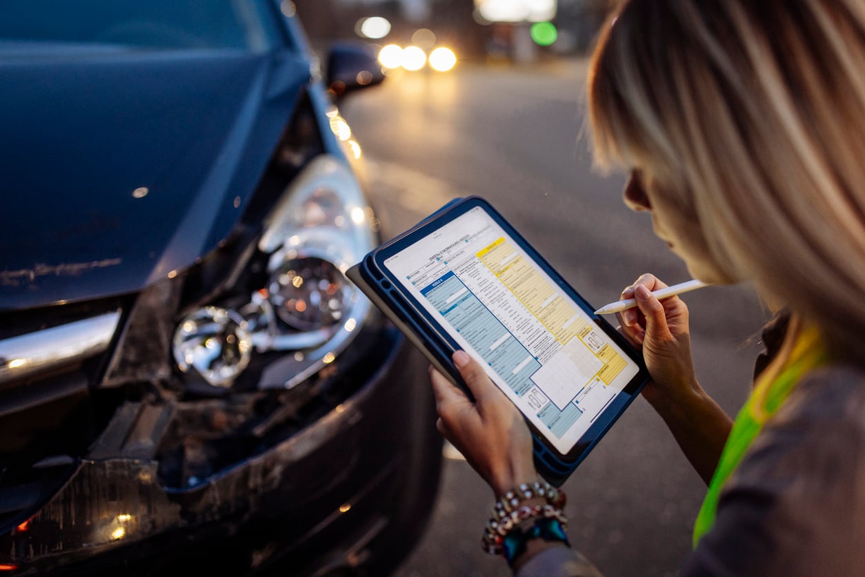 A person holding a tablet and stylus fills out an insurance form near a car with front-end damage, possibly documenting details after an accident on a city street, while considering car maintenance hacks for future prevention.