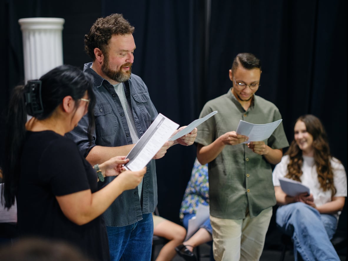 Three people stand and read scripts, smiling and interacting, while two others sit in the background, also holding papers and laughing, in what appears to be a rehearsal or group reading session—an experience reminiscent of memberships worth paying for that foster creativity and connection.