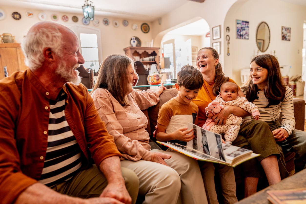 A multigenerational family sits together on a couch, smiling and laughing while looking at a photo album—a scene reminiscent of Middle-Class Moms in the 80s and 90s. A woman holds a baby, and a young boy shows photos to the group in a cozy, warmly lit living room.