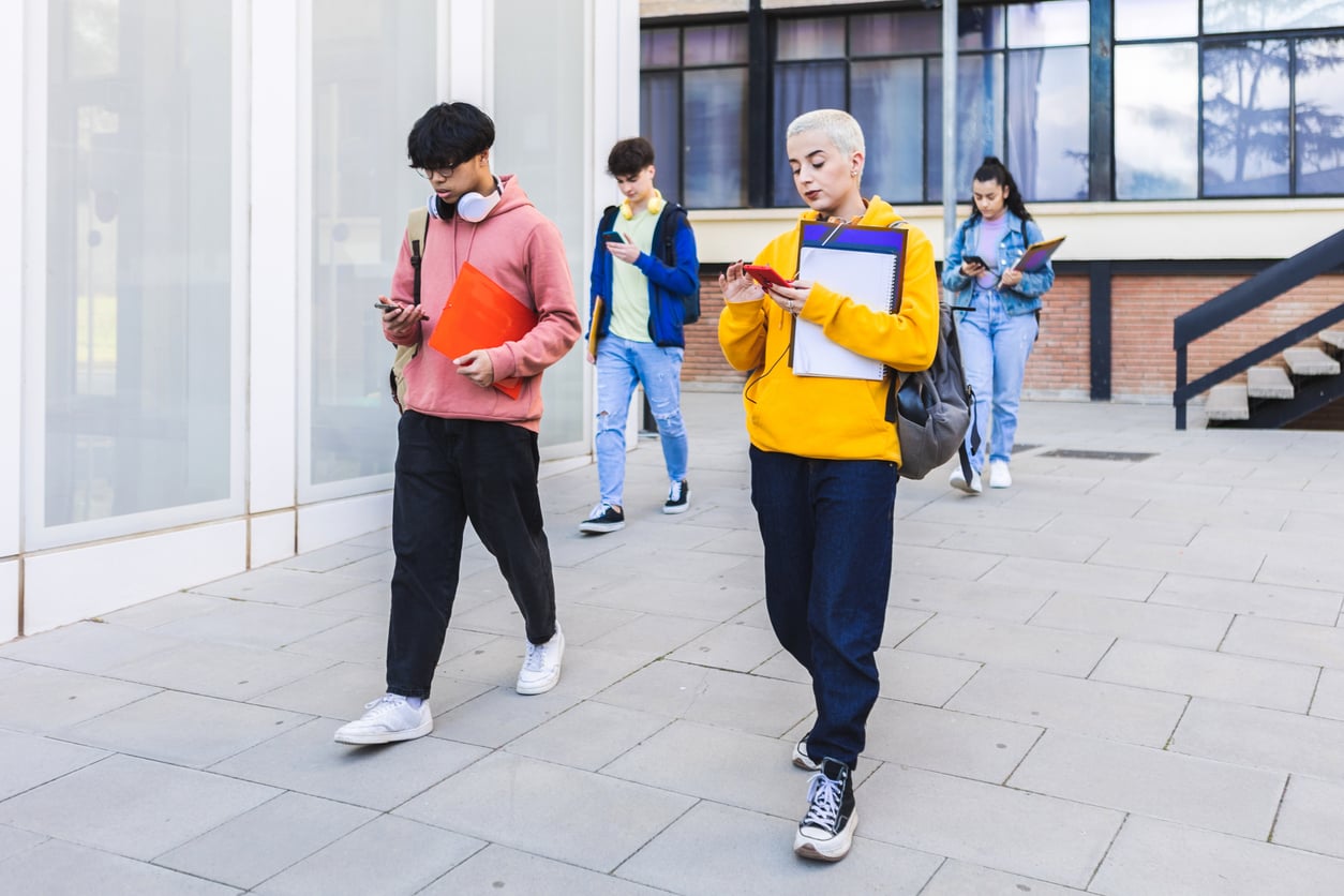 Five students walk outside a building, holding books and looking at their phones, appearing focused and engaged with their devices. The setting is urban with glass windows and concrete walkway.