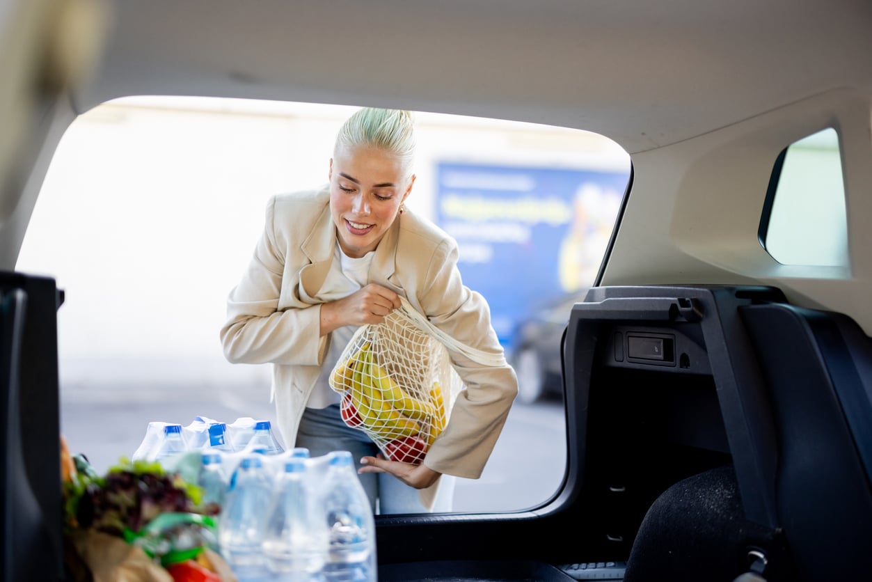 A person with light hair loads a reusable mesh bag of groceries into a car trunk filled with bottled water and produce, thoughtfully preparing their road trip checklist.