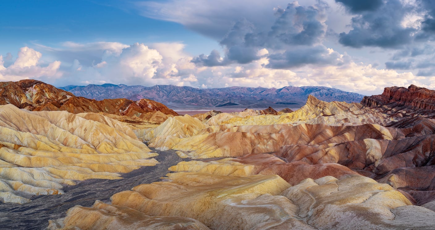 Colorful, layered rock formations under a cloudy sky in a desert landscape, with distant mountains in the background and dramatic lighting highlighting the unique terrain—one of the most stunning travel destinations on the West Coast.