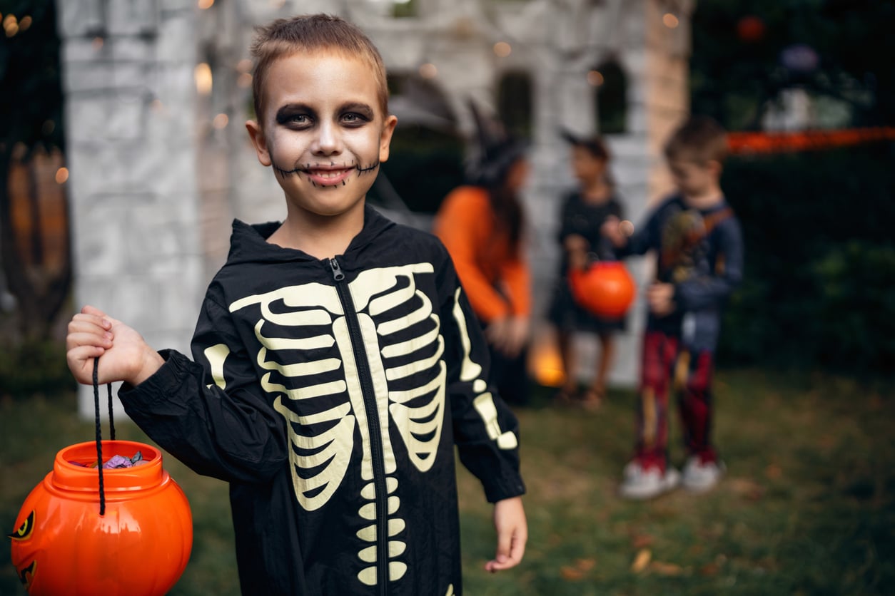 A child in a skeleton costume with face paint holds a pumpkin bucket, standing outside with other children in costumes and a spooky backdrop, ready to enjoy fun Halloween activities.
