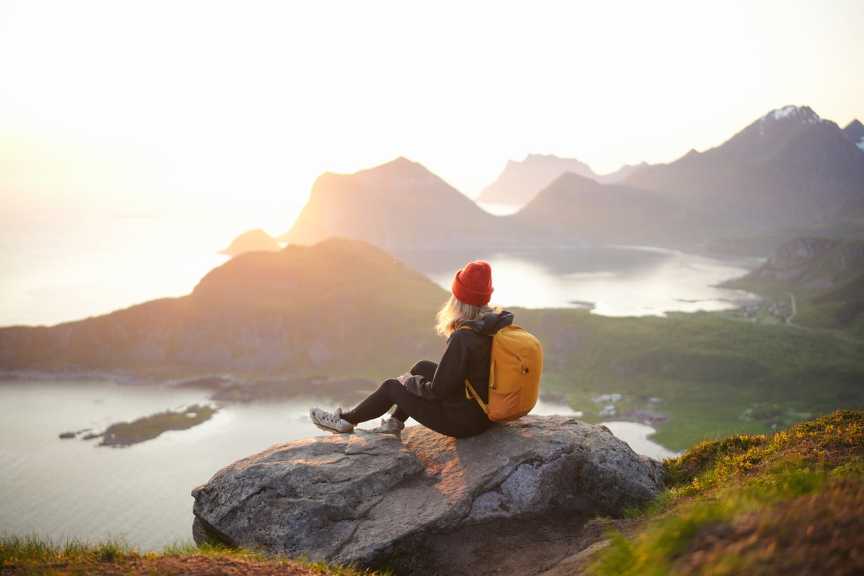 A person with a yellow backpack and red beanie sits on a rock overlooking a scenic mountain landscape at sunrise, with water and distant peaks visible, reflecting on vacation costs while taking in the breathtaking view.