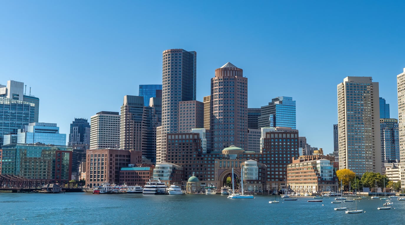 Boston city skyline with modern skyscrapers and older buildings, seen from across the harbor on a clear, sunny day. This travel-friendly scene features boats docked in the water in the foreground, inviting visitors to explore the vibrant waterfront.
