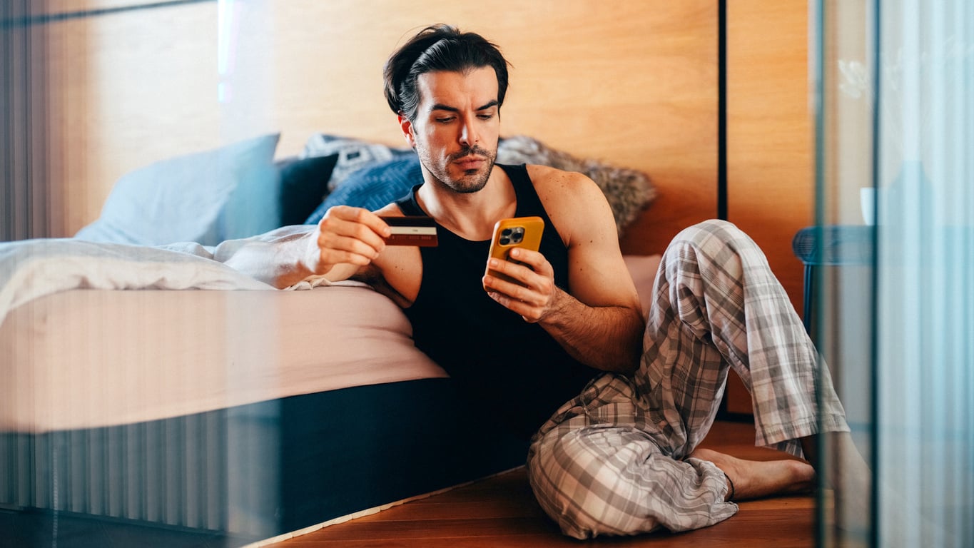 A man in pajamas sits on the floor by his bed, holding a credit card in one hand and a smartphone in the other, appearing focused on the phone screen as he practices money saving habits.