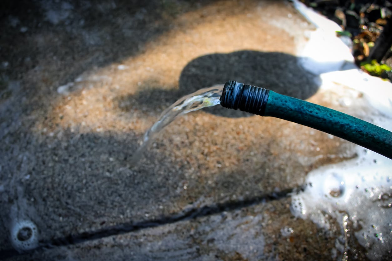 A close-up of water flowing out of a green garden hose onto a sunlit, wet concrete surface with some scattered soap suds.