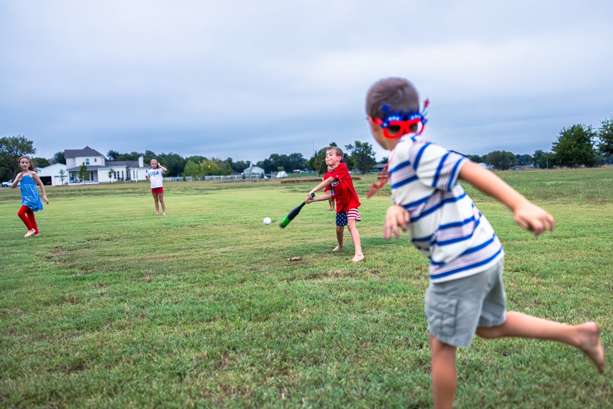 Four children play baseball on a grassy field. One child swings a bat at a ball, another pitches, and two others watch. The kids wear casual summer clothes and the sky is cloudy.