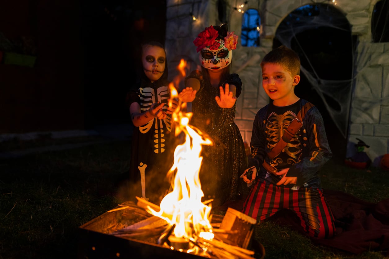 Three children in skeleton and Day of the Dead costumes sit around a small campfire at night, with a spooky, decorated house behind them, enjoying classic Halloween activities and festivities.