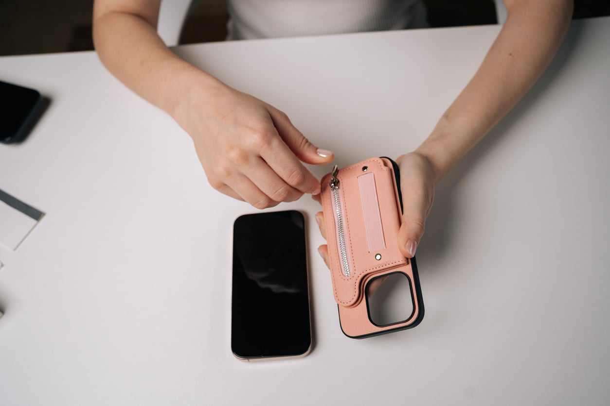 A person sits at a white desk, inserting a card into a pink phone case with a wallet slot—one of those so-called must-haves that some consider classic waste of money products. A smartphone lies face-up on the desk nearby.