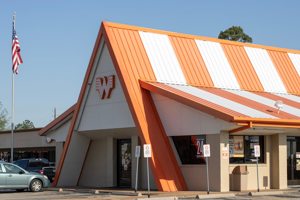 A regional fast-food chains icon, this Whataburger restaurant features an orange and white striped roof, the prominent Whataburger logo on the front, cars parked outside, and an American flag waving on a sunny day.