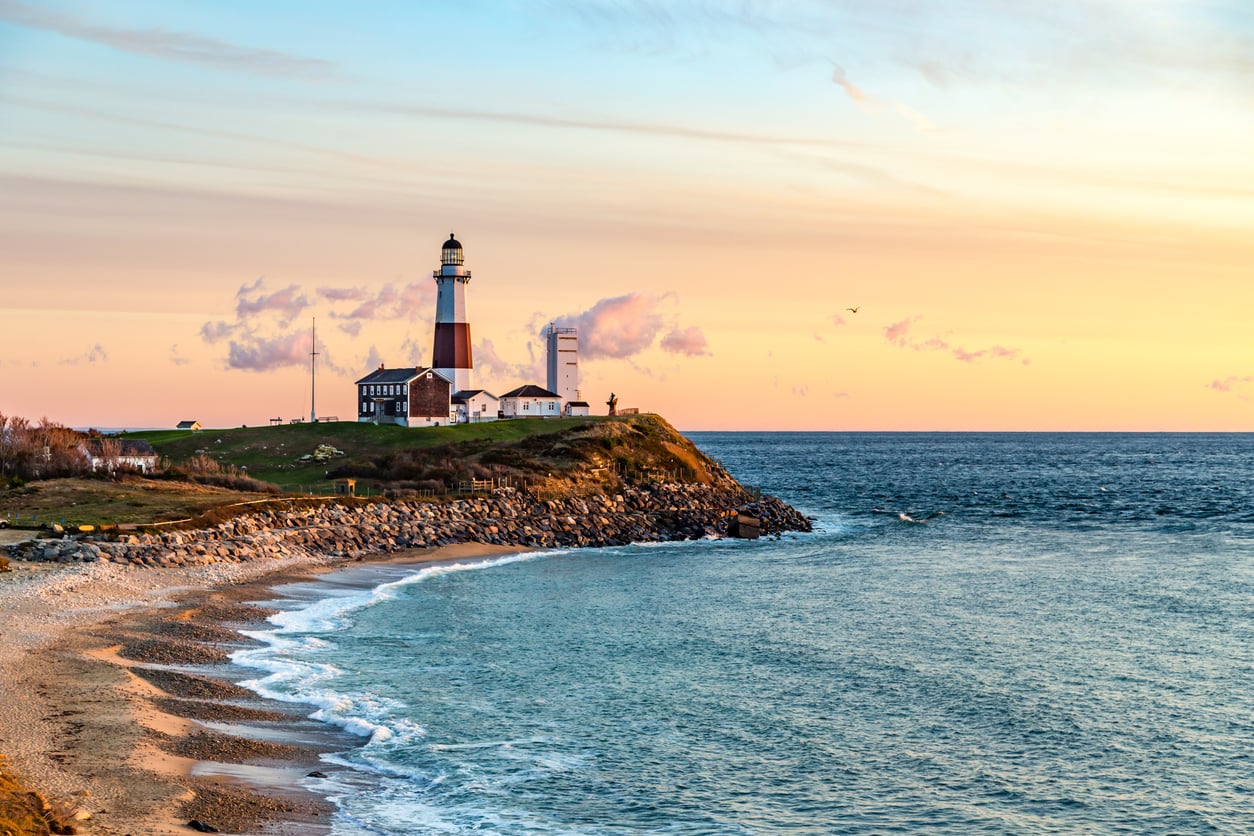 A lighthouse stands on a rocky seaside cliff at sunset, with gentle waves washing onto the shore and pastel-colored clouds in the sky.