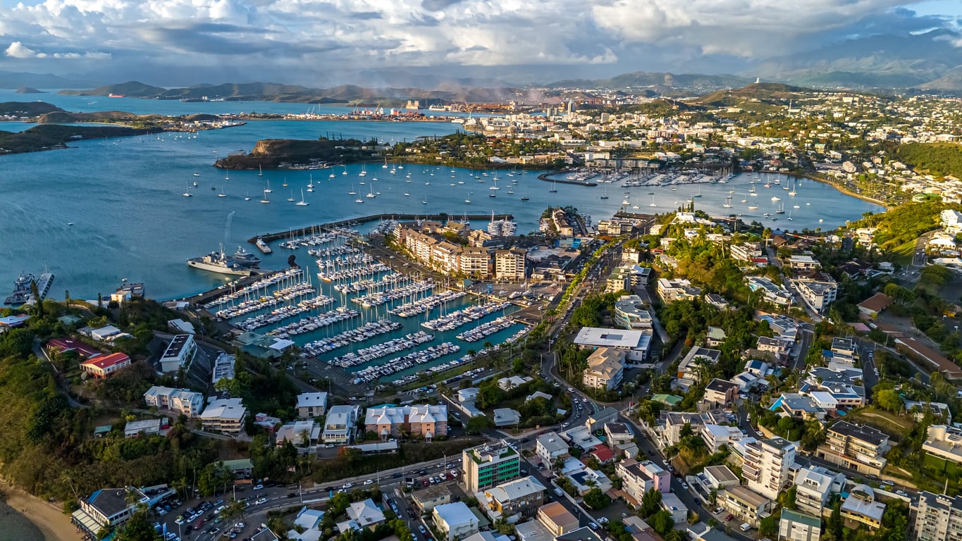 Aerial view of a coastal city, renowned among travel destinations, with a marina full of boats, surrounding buildings, green hills, and turquoise water. Islands and distant mountains are visible under partly cloudy skies.