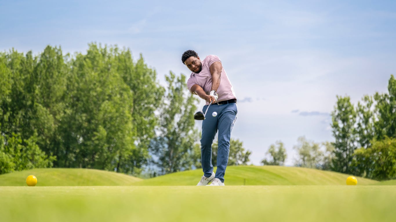 A person enjoys one of their expensive hobbies on a sunny day, swinging a golf club and hitting a golf ball from a tee, with green grass and trees in the background.
