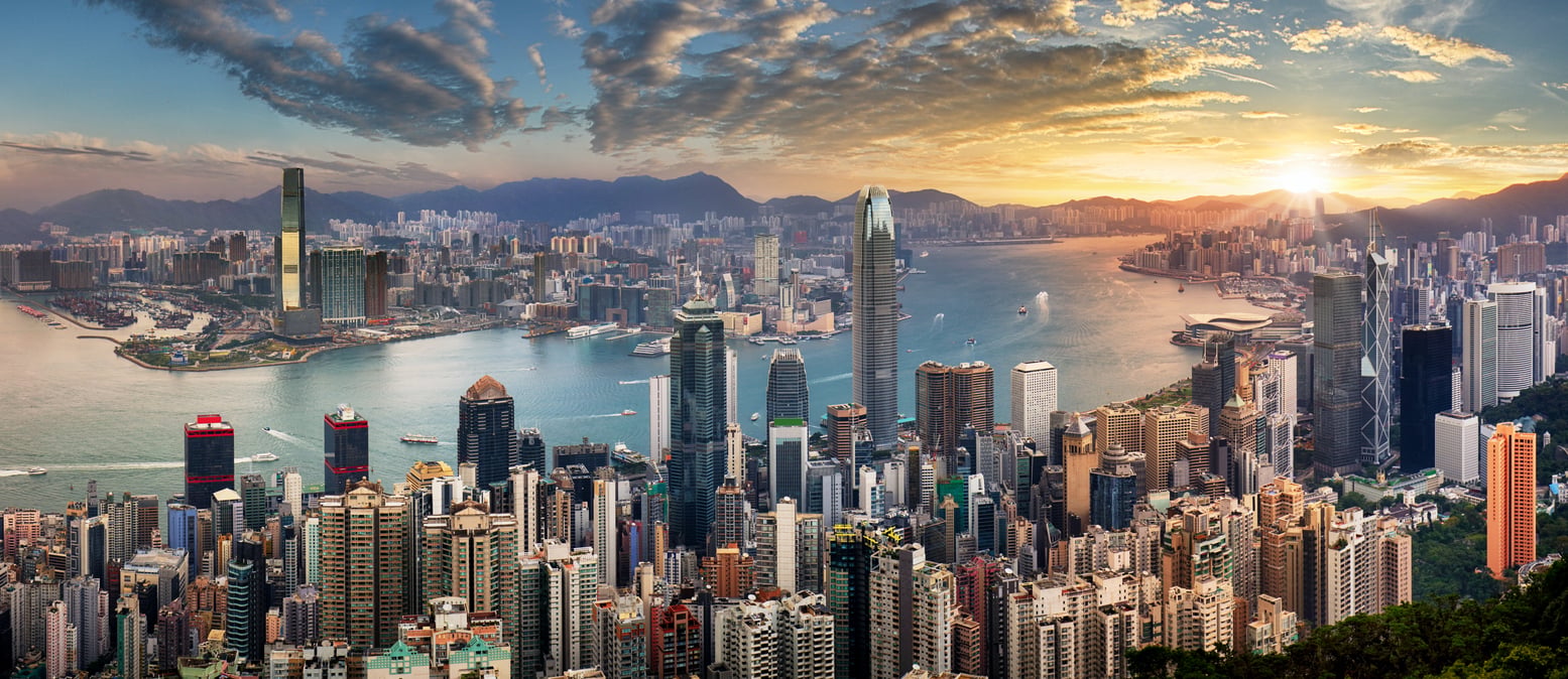 Panoramic view of a city skyline with tall skyscrapers, a harbor, and mountains in the background at sunset under a partly cloudy sky—a breathtaking scene that highlights why this location is among the world’s top travel destinations.