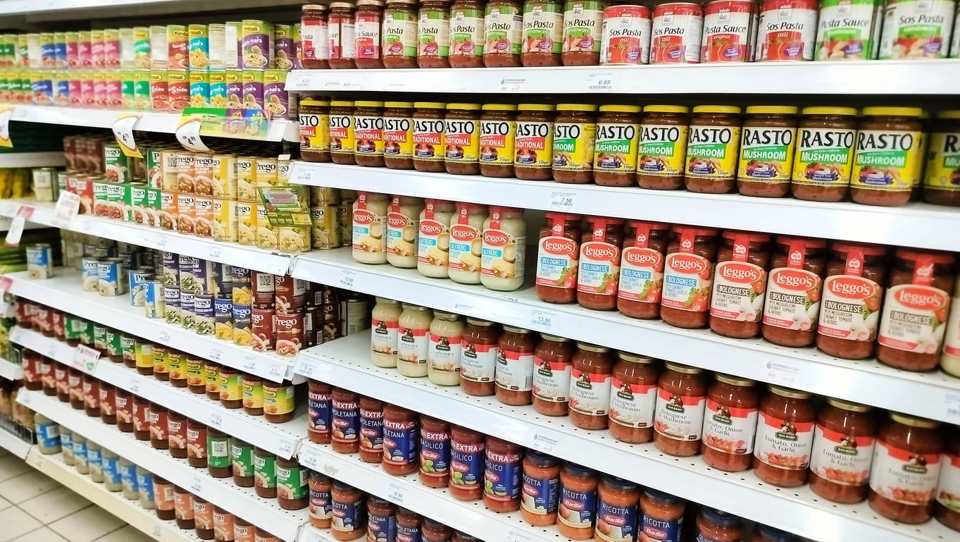 Grocery store shelves stocked with various brands and types of pasta sauce jars, canned foods, and packaged pasta boxes, organized in neat rows, reflect typical grocery shopping habits as customers seek convenience and variety during their visits.