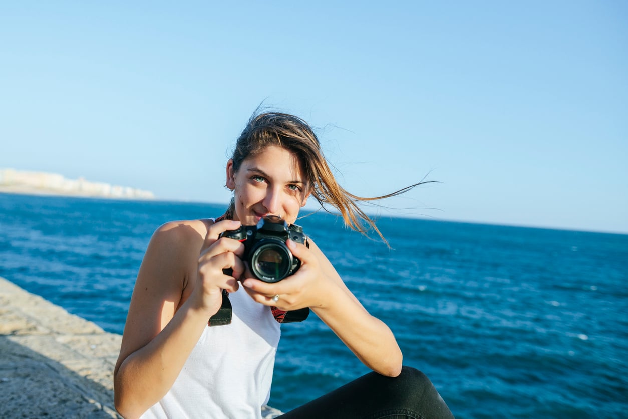 A woman with long hair holds a camera and smiles while sitting by the ocean on a sunny day, enjoying one of her expensive hobbies. The sky and water are blue, and there is a stone wall next to her.