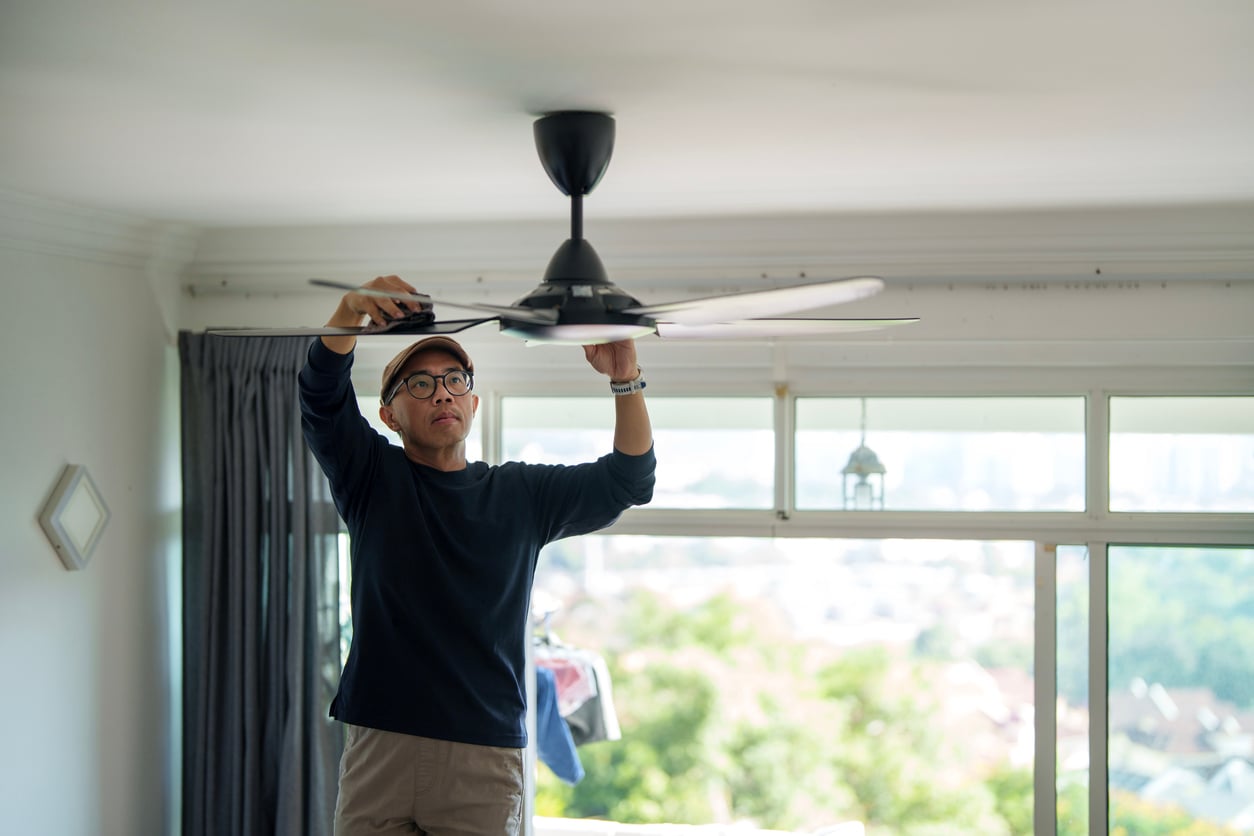 A man wearing glasses and a cap stands on the floor and cleans or repairs a ceiling fan in a bright room with large windows and curtains.