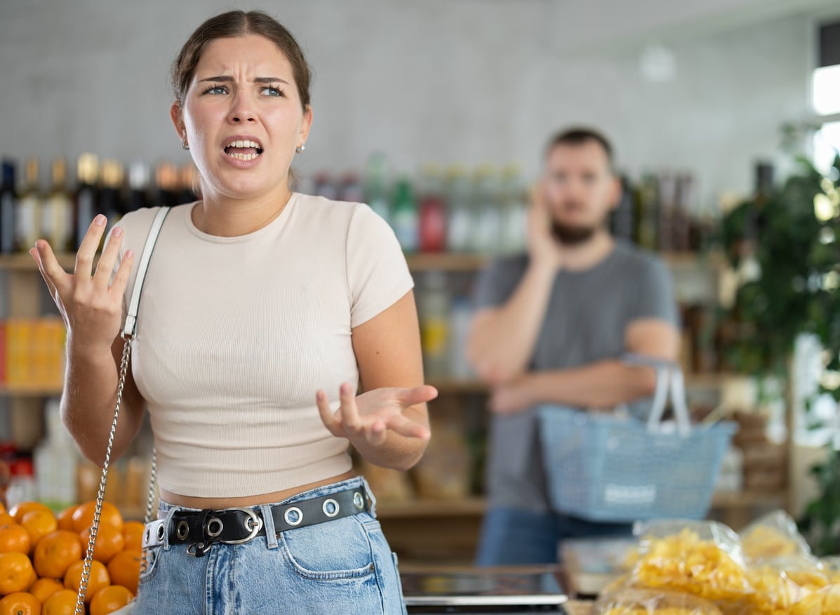 A young woman in a grocery store looks frustrated, gesturing with one hand, while a man in the background stands with a shopping basket, appearing concerned—perhaps overwhelmed by the abundance of fast-food grocery store products on the shelves.