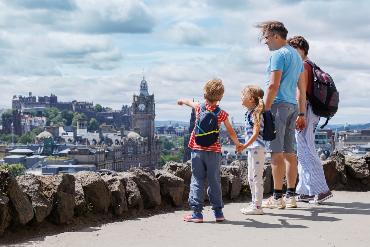 A family with two children looks out over a cityscape from a high viewpoint; the boy points towards historic buildings under a partly cloudy sky, perhaps imagining the adventures made possible through vacation swaps.