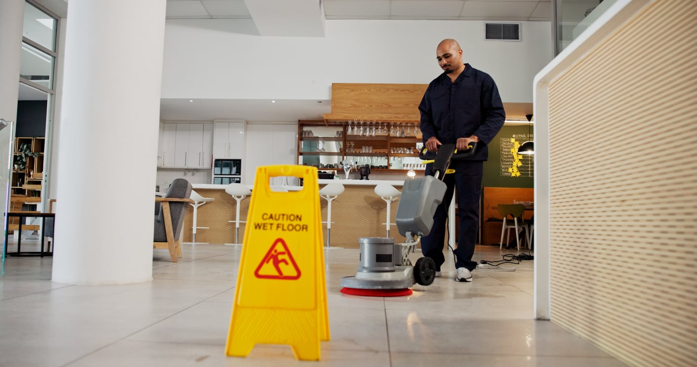 A worker in dark clothing uses a floor-cleaning machine in a modern, bright room, demonstrating the Baby Boomer beliefs in hard work and attention to detail. A yellow “Caution Wet Floor” sign is placed prominently in the foreground on the tile.