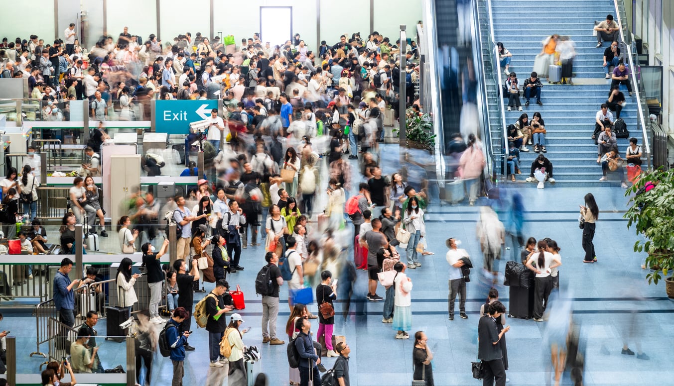 A large, busy crowd fills an indoor space, with people walking, standing, and waiting. Some are on escalators or sitting on steps. Blue flooring and an “Exit” sign are visible. The scene is lively and bustling.