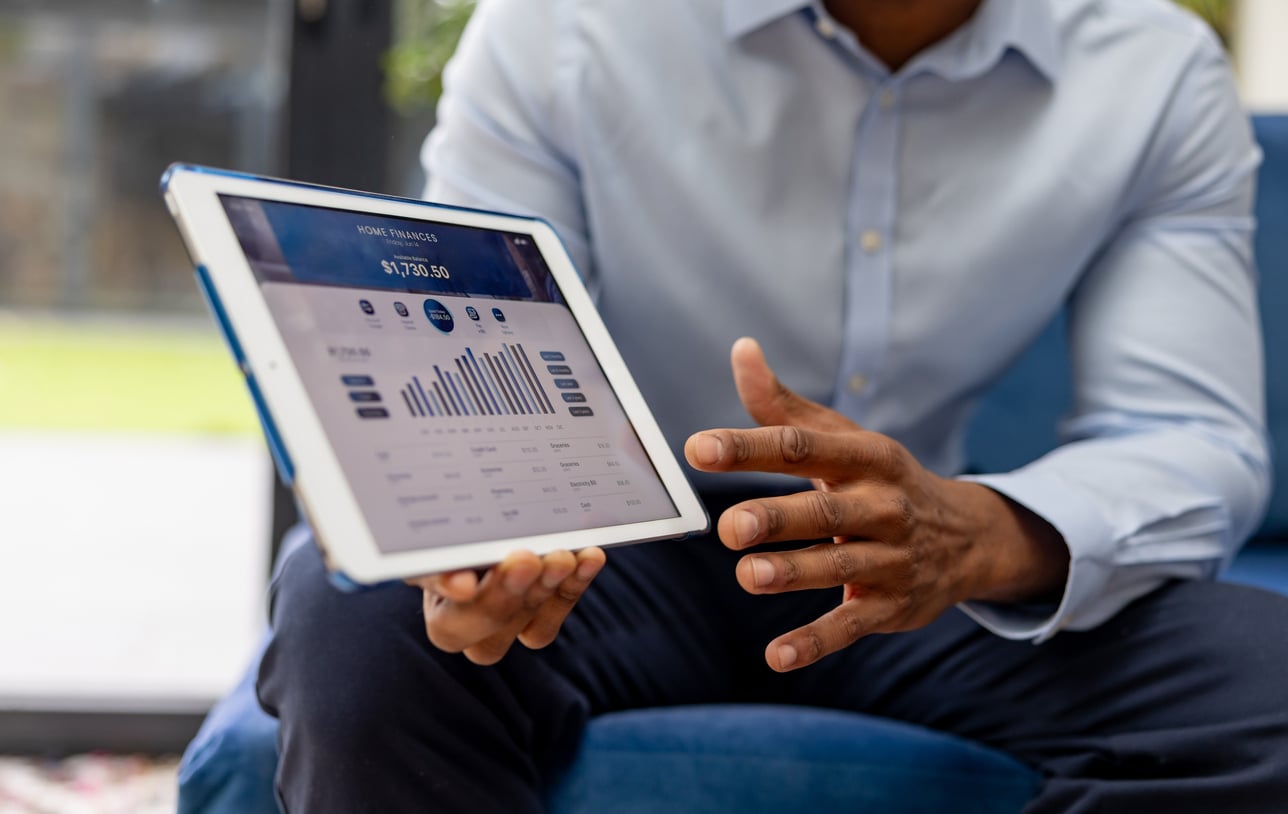 A person in a light blue shirt holds a tablet displaying financial charts and graphs on household expenses, presenting data with one hand while sitting on a blue chair.