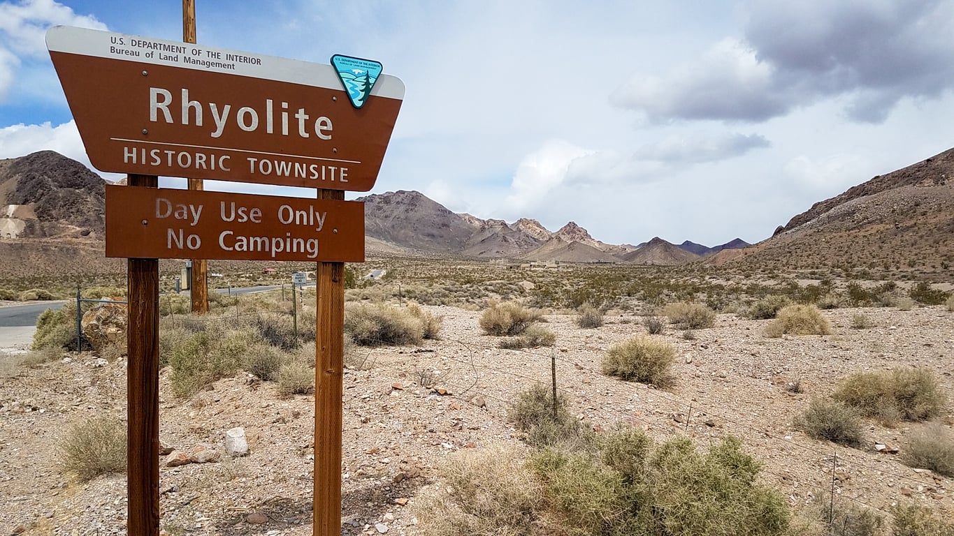 A wooden sign reads "Rhyolite Historic Townsite. Day Use Only. No Camping" in a desert landscape—a reminder of the fascinating legacy found in many American towns—with mountains rising in the background under a partly cloudy sky.