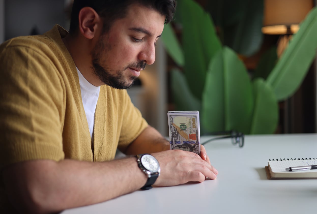 A man in a yellow sweater sits at a table, holding a stack of hundred-dollar bills—perhaps contemplating his money saving habits. A notebook and pen are on the table, ready for budgeting, while large green plant leaves provide a calming backdrop.