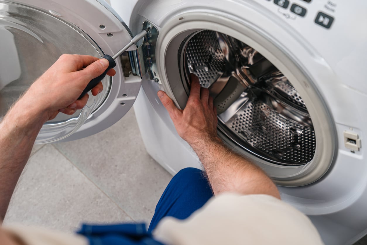 A person uses a screwdriver to repair the inside panel of a washing machine door, highlighting the frustration of products that don't last, with the drum visible and the machine open.