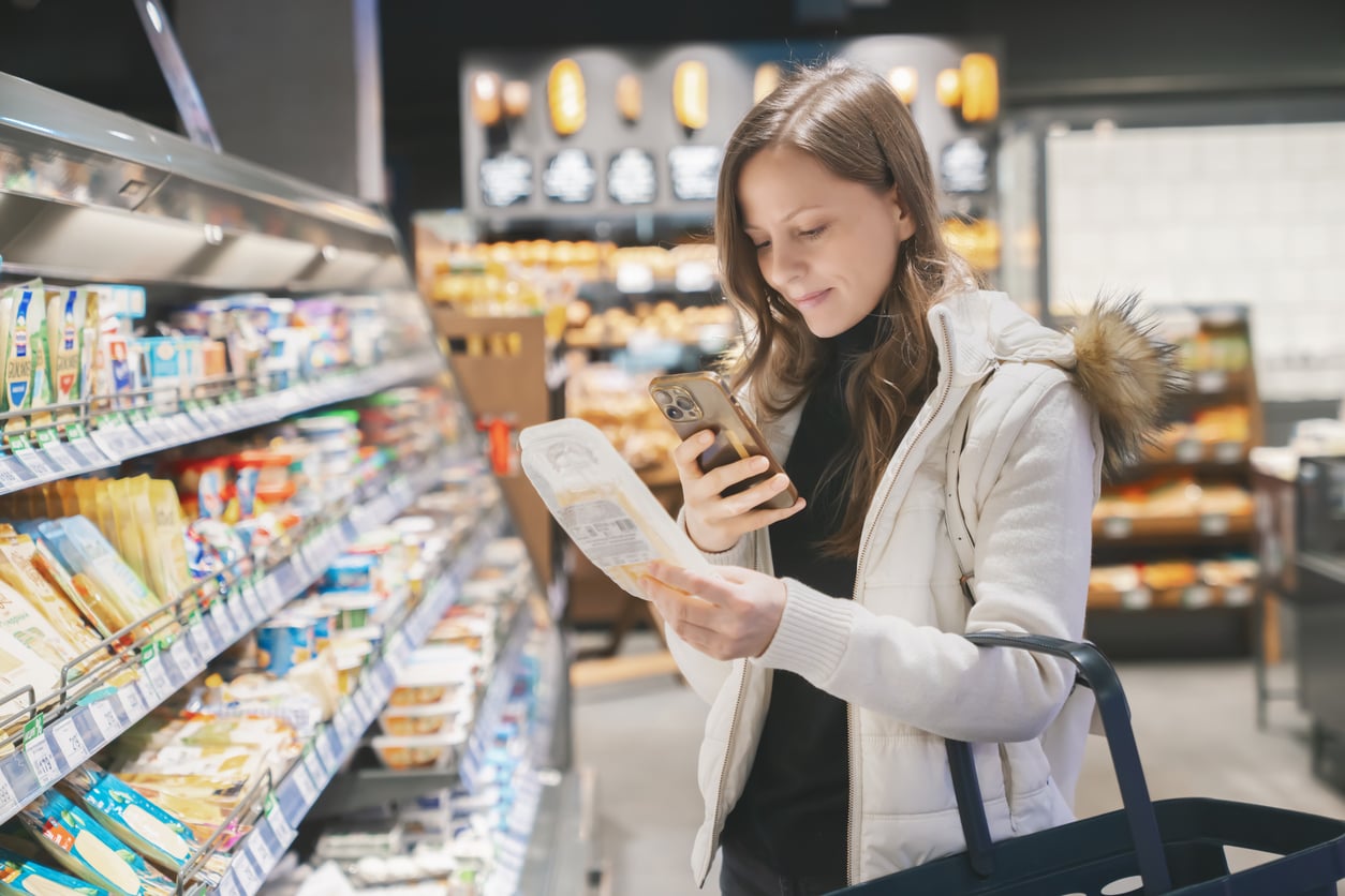 A woman in a white jacket holds a shopping basket and scans a food package with her phone in a grocery store aisle filled with dairy products, reflecting modern grocery shopping habits.