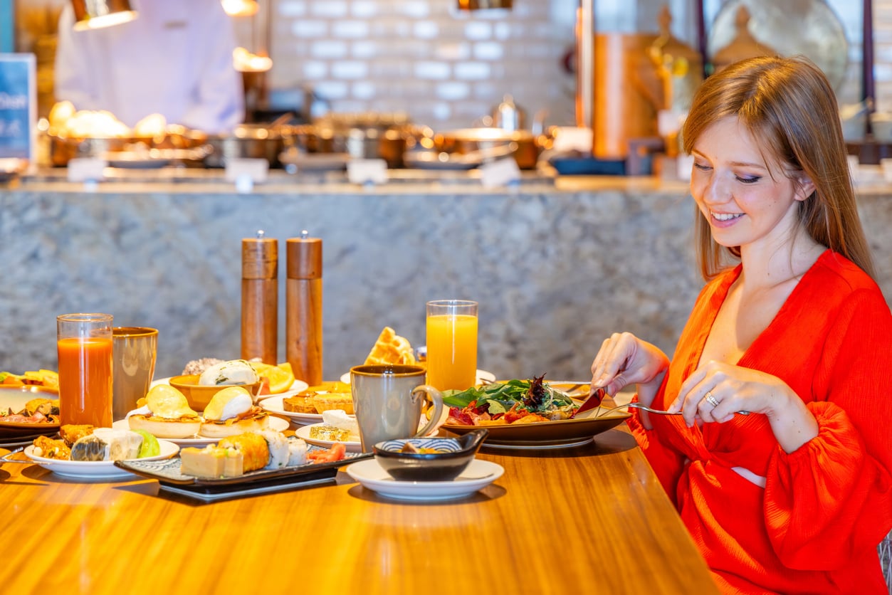 A woman in a red dress smiles while dining alone at a restaurant table, which is filled with various breakfast foods, pastries, and drinks, in a bright, modern setting—perhaps savoring a moment of indulgence without worrying about vacation costs.
