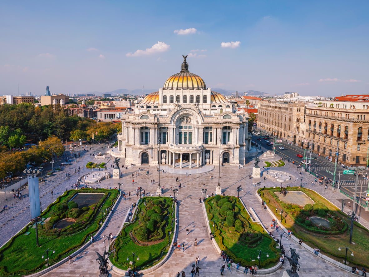 Aerial view of Palacio de Bellas Artes in Mexico City, one of the city's must-see destinations, with its domed roof, ornate facade, surrounding gardens, and people walking around on a sunny day.