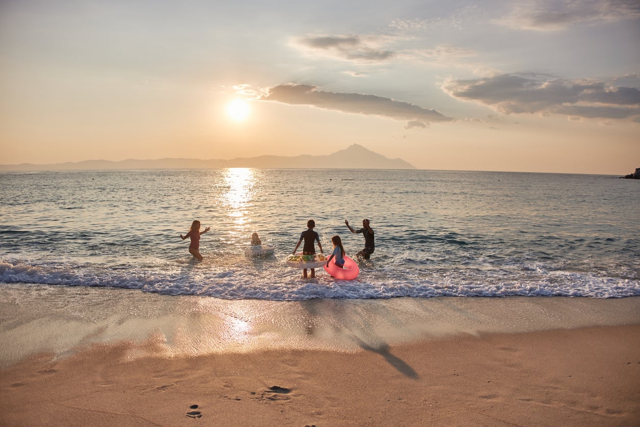 Five people play in the ocean at sunset, with gentle waves and mountains in the background. Enjoying a moment made possible by vacation swaps, one person holds a pink inflatable ring, and their shadows stretch across the sandy beach.