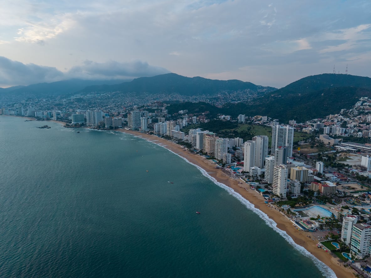 Aerial view of a coastal city with tall buildings along a sandy beach, waves hitting the shore, and green mountains in the background under a cloudy sky, making it one of the most captivating travel destinations to explore.