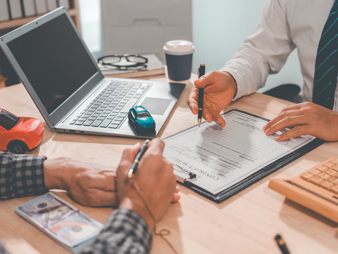 Two people sit at a desk, one signing a contract form. A laptop, toy cars, coffee cup, and cash are on the table, indicating a car purchase or insurance agreement as part of managing household expenses.