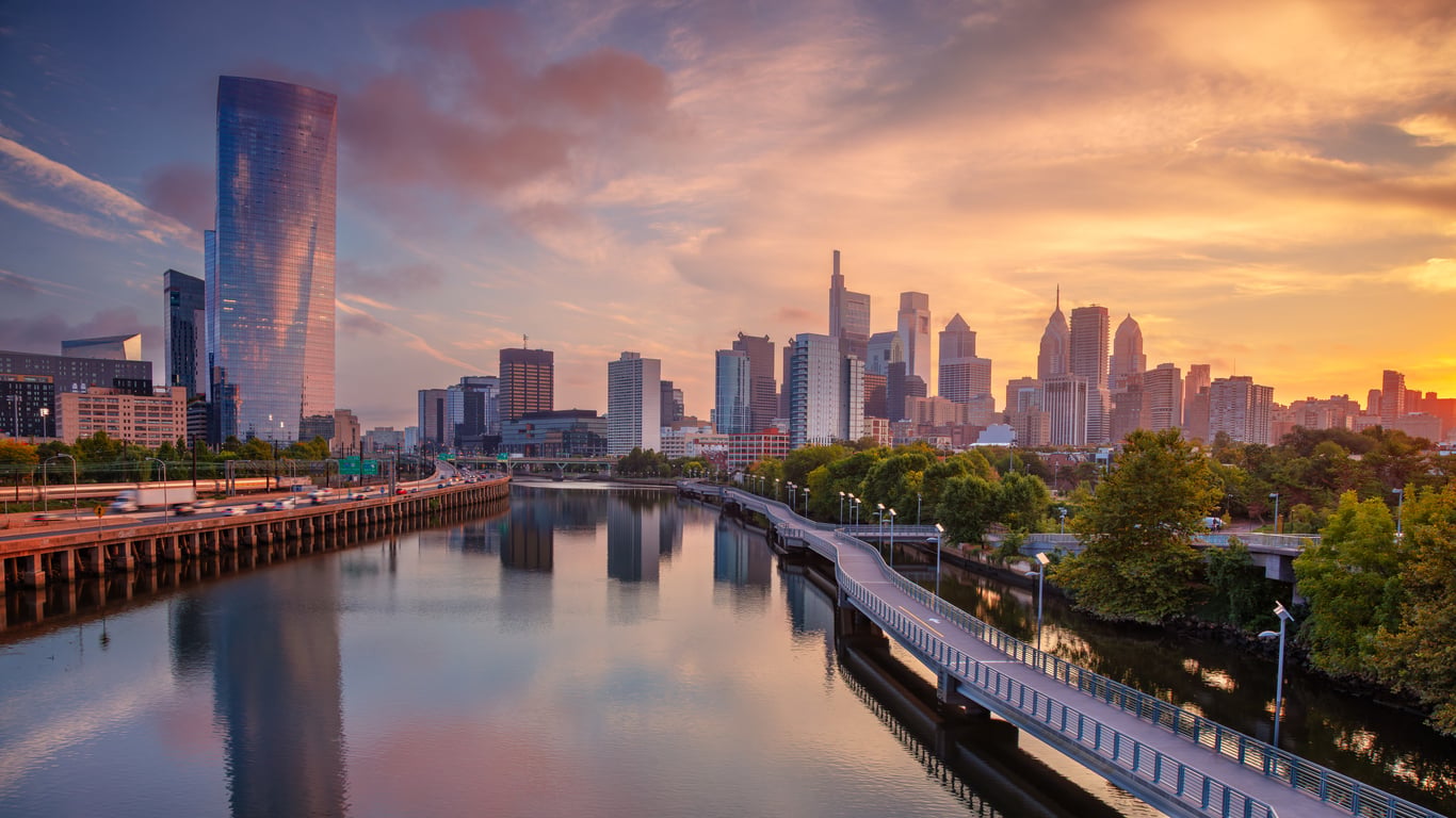 A city skyline at sunset reflects on a river, with modern skyscrapers reminiscent of big cities in America, a curving pedestrian walkway, and lush green trees along the water’s edge under a colorful sky.