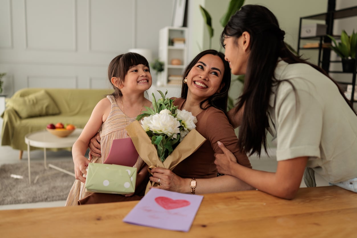 A woman smiles while holding a bouquet of white flowers, seated beside a young girl with a gift and a card. Another young woman stands nearby, smiling. A handmade card with a heart is on the table in front of them.