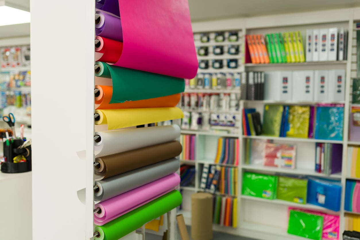 Rolls of colorful wrapping paper are displayed vertically on a stand in a Walmart store, with shelves of stationery items, binders, and paper products visible in the background.
