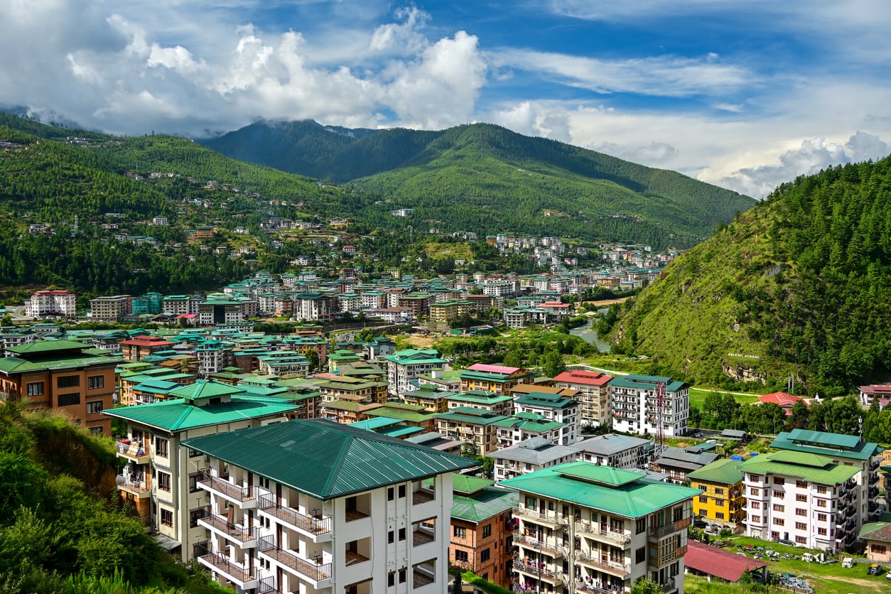 A city with colorful buildings and green roofs nestled among lush hills and mountains under a partly cloudy sky, making it one of the most enchanting travel destinations for those seeking scenic beauty.