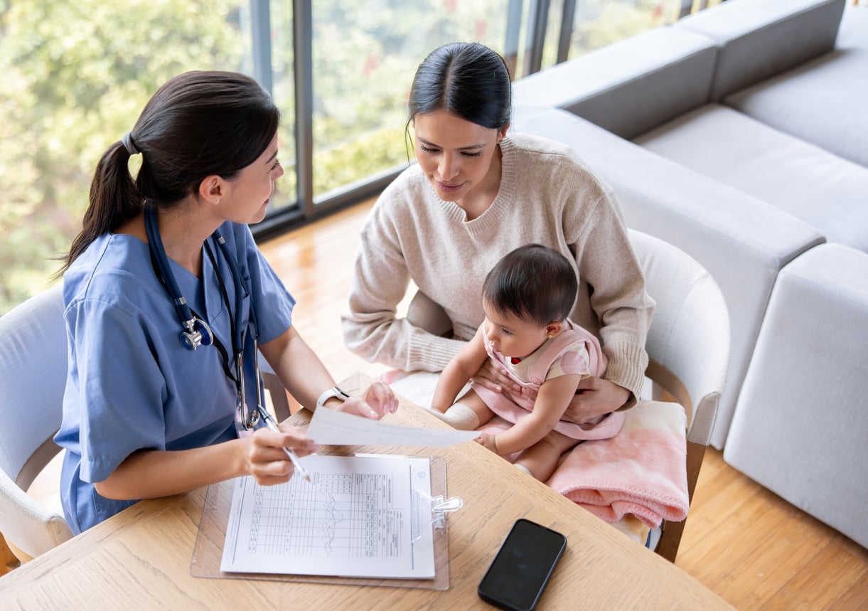 A doctor in blue scrubs discusses paperwork with a woman holding a baby on her lap in a bright, modern room, reflecting how families in America are having fewer children. Medical charts and a smartphone are on the table.