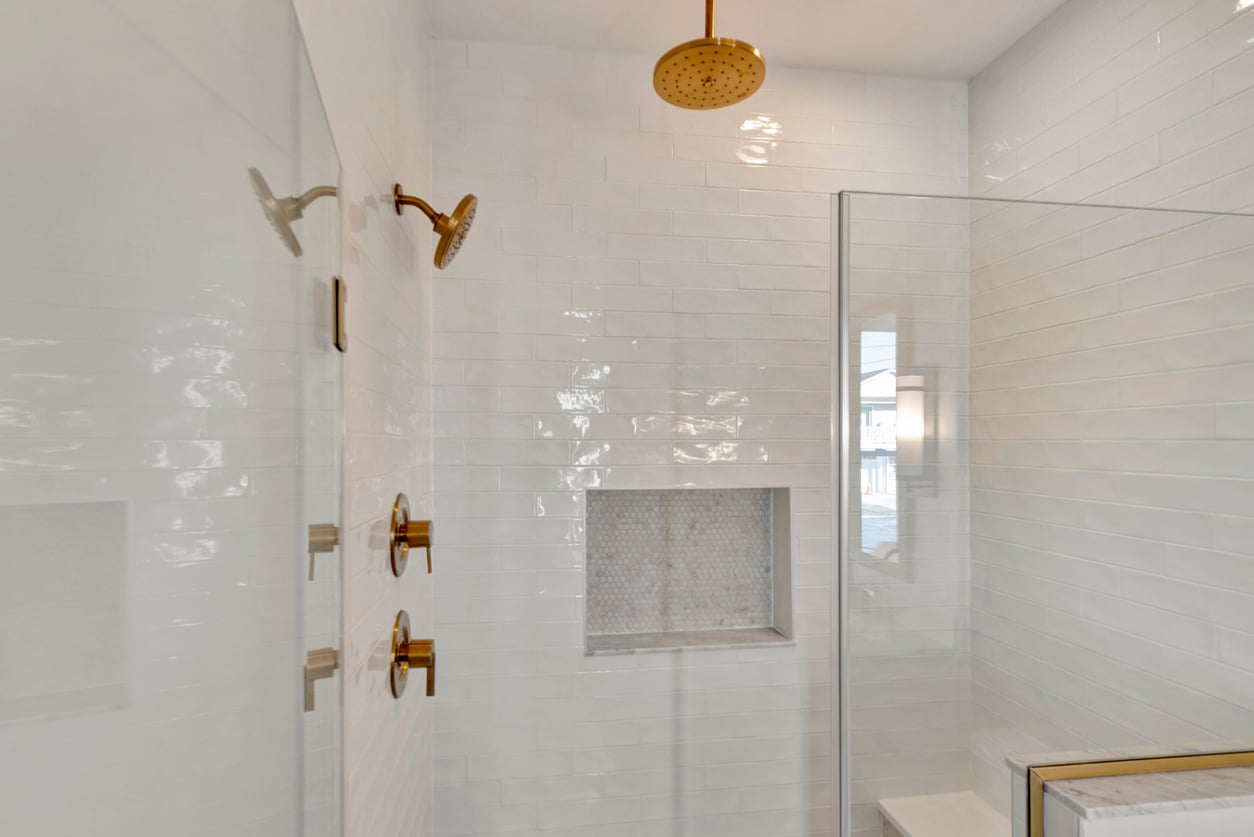 Home features. Modern shower with white subway tile walls, gold fixtures including a rainfall showerhead and two standard showerheads, built-in recessed shelf with mosaic tile, and a glass enclosure.