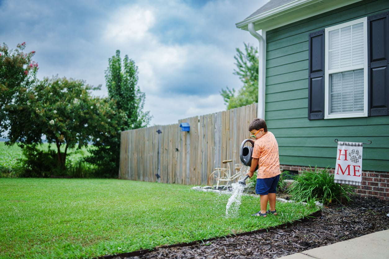 A young boy waters the lawn with a hose beside a green house on a cloudy day. A "HOME" sign hangs near the house, and a wooden fence and trees are in the background.