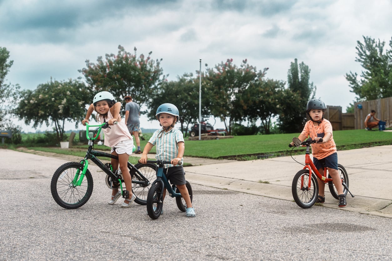 Three young children wearing helmets are riding bicycles on a paved street. Trees and greenery are visible in the background, and an adult walks nearby under a cloudy sky.